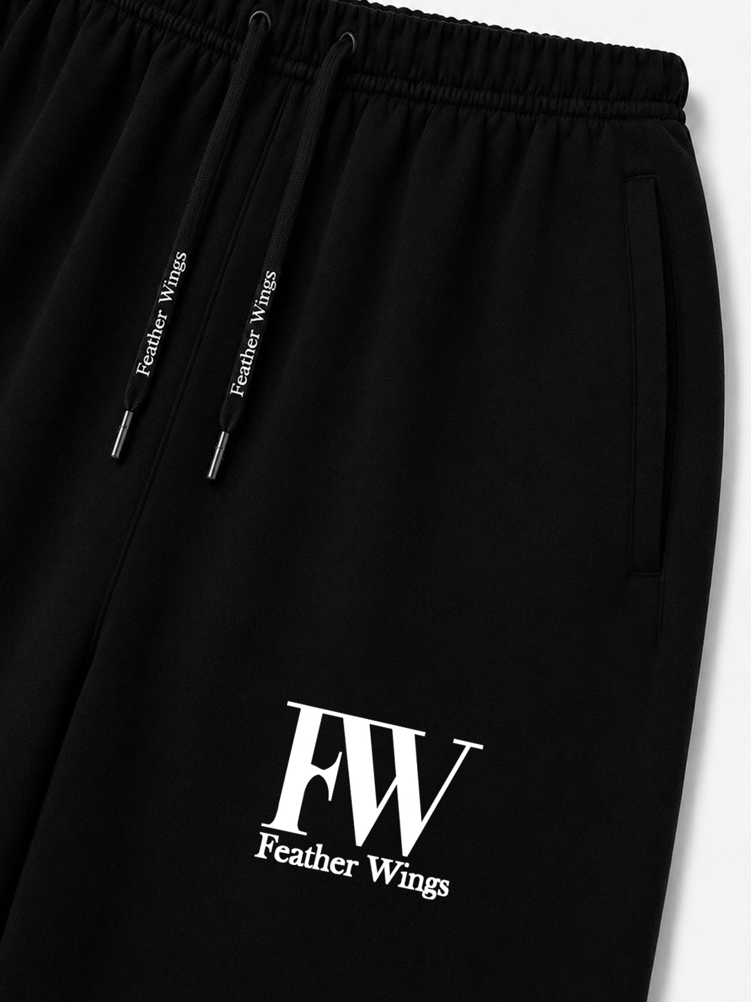 Close-up of black drawstring sweatpants with a white 'FW Feather Wings' logo printed near the lower left leg, shown against a light gray background.