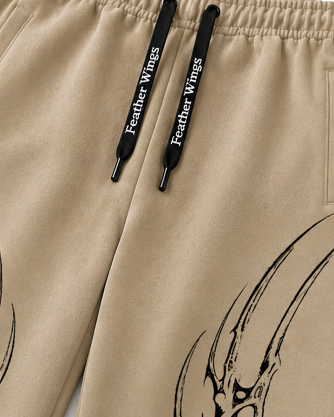 Close-up of beige drawstring pants with black drawstrings printed with 'Feather Wings' and a black graphic design on the leg.
